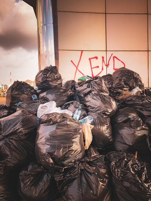A large pile of black plastic rubbish bags, some tied and others loosely closed, stacked against a modern building with smooth, beige-colored tiled exterior panels. The bags appear to contain mixed wa