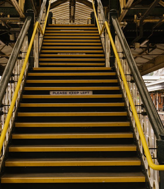 A straight flight of industrial stairs with yellow safety risers and black anti-slip treads, situated within a warehouse or factory environment. The staircase is flanked by metal handrails painted yel